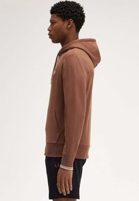 Felpa con cappuccio marrone, dotata di tasca frontale, polsini a coste e cordino. Indossata sopra pantaloni corti neri con logo ricamato sulla coscia sinistra.