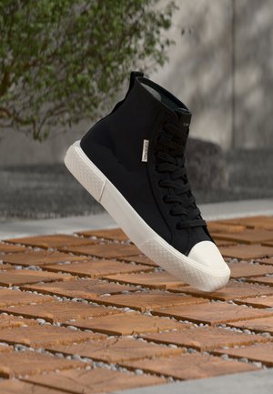 Zapatilla negra de caña alta con suela blanca y puntera blanca, flotando sobre baldosas de madera con pequeñas piedras grises entre ellas.