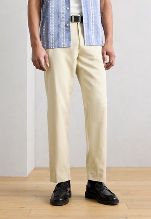 Άνδρας που φοράει μπεζ παντελόνι, μαύρη ζώνη, μαύρα loafers και μπλε πουκάμισο με κοντά μανίκια και σχέδιο πάνω από λευκό εσώρουχο, στέκεται σε ξύλινο δάπεδο.