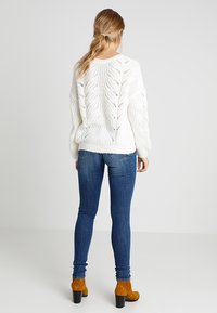 Femme aux cheveux blonds attachés en queue de cheval basse, portant un pull blanc en maille torsadée, un jean skinny bleu et des bottines en daim moutarde à talons carrés.