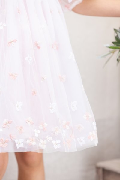 Une robe en tulle rose ornée de motifs floraux brodés en blanc et pêche, avec une texture douce et semi-transparente et une silhouette fluide.