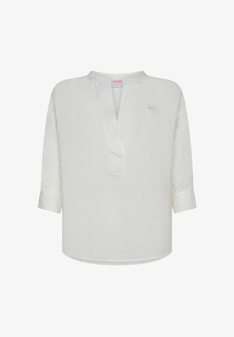 Camicia tunica bianca in lino con scollatura a V, maniche lunghe e orlo leggermente curvo. Presenta un piccolo logo dorato sul lato sinistro.