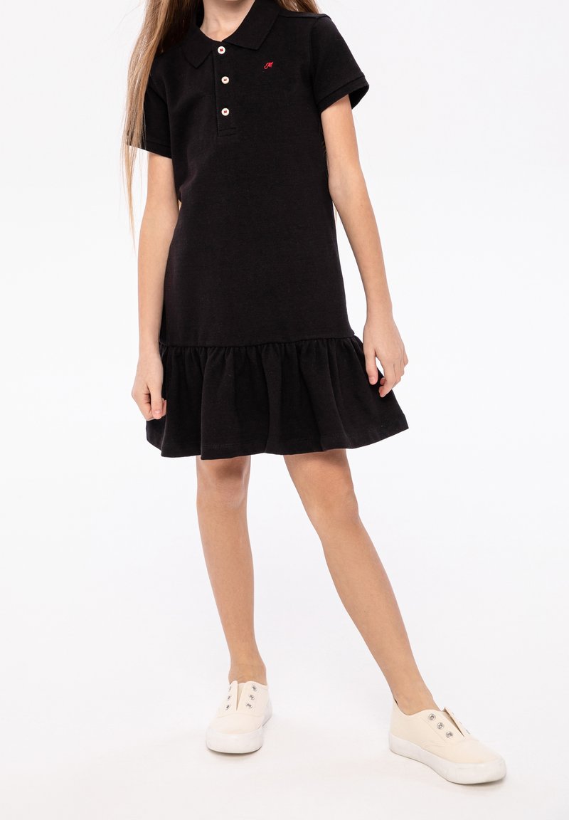 Fille portant une robe polo noire à manches courtes avec des boutons et des baskets blanches sans lacets, debout devant un fond blanc uni.