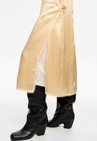 Abito beige seta con spacco laterale e bordo in pizzo, abbinato a stivali neri in pelle al ginocchio con tacco alto.