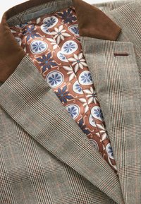 Brunternet blazer med en blød brun krave og et farverigt blomster-mønsteret indvendigt foer i blå, hvide og brune nuancer.