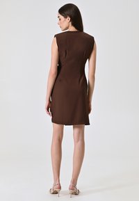 Braunes, ärmelloses Kleid mit taillierter Passform und Rückenabnähern. Glattes Material mit einem knielangen Saum. Das Modell trägt beige High-Heels.