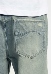 Jeans di denim azzurro chiaro con tasca posteriore leggermente rovinata nell'angolo superiore, indossati con una camicia bianca infilata dentro.
