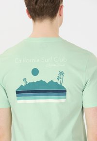 Vihreä mintunvärinen puuvillapaita, jossa on graafinen kuva vuorista ja palmupuista, sekä sinisistä ja valkoisista kerrostetuista vaakasuorista raidoista. Teksti lukee "California Surf Club Malibu Beach."