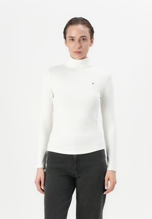 Maglione bianco a collo alto con maniche lunghe e design aderente. Presenta un piccolo logo vicino al petto ed è abbinato a pantaloni grigio scuro.