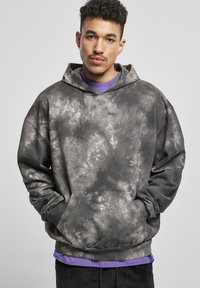 Sivi hoodie s uzorkom od batik boje s prednjim džepom, velikom kapom i rebrastim manžetama, složen preko ljubičaste majice, prikazan na neutralnoj pozadini.