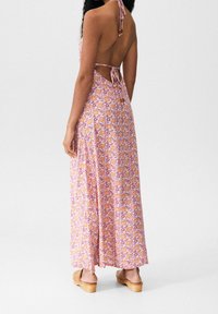 Vestido largo floral con fondo rosa, patrones de flores amarillas y blancas, diseño de espalda descubierta y lazos ajustables. Combinado con sandalias de plataforma de madera.