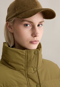 Olijfgroene pufferjack met hoge kraag en drukknoopsluiting, gecombineerd met een textuurrijk bruin corduroy honkbalpet. Neutrale achtergrond.