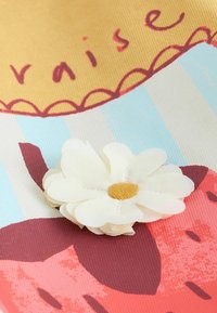 Floare albă din material textil cu centru galben pe o suprafață colorată ce prezintă căpșuni roșii și dungi albastre, cu text și margini decorative zimțate.