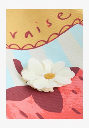 Biały materiałowy kwiat z żółtym środkiem na kolorowej powierzchni z czerwonymi truskawkami i niebieskimi paskami, z tekstem i ozdobnymi karbowanymi krawędziami.