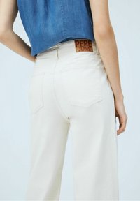 Beige leveleiset housut, jotka ovat denimistä valmistetut, takataskuilla ja punaisella "Pepe Jeans" -logolla vyötäröllä, yhdistetty siniseen yläosaan.