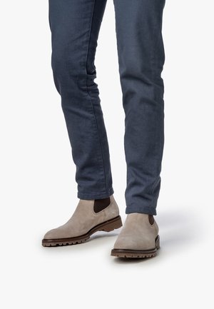 Beige suède enklaarzen met elastische zijpanelen, een bruine rubberen zool en een gestructureerde afwerking, gedragen met donkerblauwe slim-fit broek.