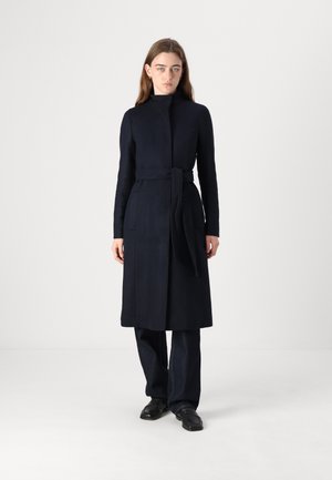 Marineblå frakke med høj krave, bæltestrop i taljen, lange ærmer og sidelommer. Båret over mørke denimbukser og sorte sko.
