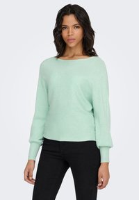 Maglione verde menta a coste con scollo ampio e maniche lunghe leggermente gonfie, abbinato a pantaloni neri aderenti. La texture sembra morbida.