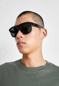 Ochelari de soare rectangulari negri cu lentile închise, având o formă distinctivă și branding pe templu. Purtați de o persoană într-o cămașă verde.