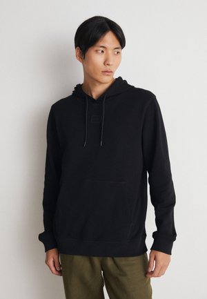 Haut à capuche en coton noir avec une poche avant, un capuchon à cordon et une étiquette logo sur la poitrine. Présente des poignets et un ourlet côtelés. Porté avec un pantalon vert olive.