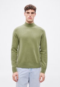 Zöld gyapjúpulóver magas nyakkal, bordázott mandzsettákkal és szegéllyel; laza szabású és enyhe textúrájú, világoskék farmerral párosítva.