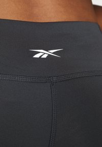 Černé atletické legíny s hladkou texturou, s bílým logem blízko pasu a kontrastním prošitím na švech.