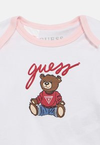 Λευκό βρεφικό κορμάκι με ροζ περιθώριο που απεικονίζει ένα καφέ αρκουδάκι με κόκκινο πουλόβερ και μπλε τζιν, με την επιγραφή «guess» από πάνω.