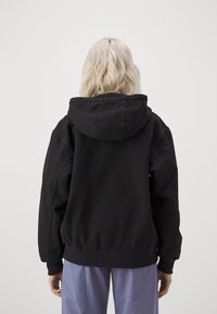 Black hooded sweatshirt with a relaxed fit, fleece texture, ribbed cuffs, and hem. Features a large hood and is worn over light trousers.