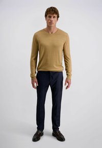 Man staat in een beige lange mouw shirt, donkerblauwe broek en donkerbruine instappers voor een effen lichte achtergrond.