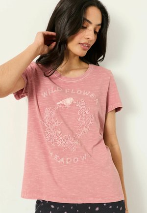 Młoda kobieta o ciemnych włosach, ubrana w różowy t-shirt z wzorem ptaka i kwiatowego wieńca oraz napisem wildflower meadow.