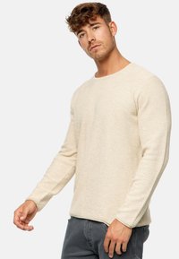 Helles beiges Langarmpullover mit strukturierter Strickmuster, rundem Ausschnitt und geradem Saum, kombiniert mit dunkelgrauen Jeans.