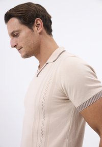 Beige kortærmet polo t-shirt med V-hals, som har teksturerede lodrette striber og en kontrasterende brun krave og manchetter.