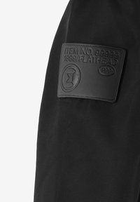 Czarna tkanina z teksturowaną skórzaną łatą z wypukłymi detalami: "ITEM NO. 99999 1999/FLATHEAD PPP" oraz okrągłym elementem dekoracyjnym.