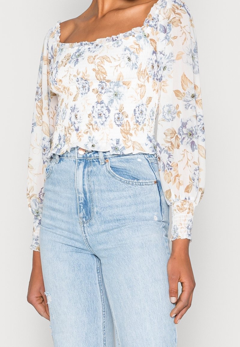 Blusa floreale con maniche lunghe a palloncino, caratterizzata da un collo a scomparsa, colori blu chiaro e crema, e una texture gather. Abbinata a jeans in denim chiaro.