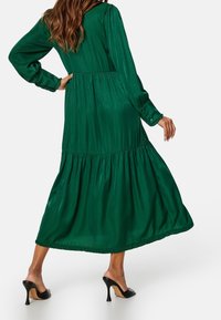 Grön, långärmad, lager-på-lager maxiklänning gjord av glatt tyg med en sammanknuten midjedesign. Har en volangkant och knäppbara manschetter.