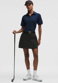 Camisa polo marina de manga corta, falda de golf negra, cinturón negro, zapatos de golf blancos y una gorra negra, de pie con un palo de golf.