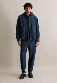 Jeansjacke mit Kapuze, zwei vorderen Taschen, kombiniert mit passenden Jeanshosen. Dunkelblaue Farbe, lockere Passform, mit sichtbaren Nahtdetails.