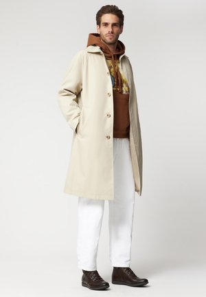 Beżowy trencz o prostym kroju, z dużymi guzami i bocznymi kieszeniami. Noszony na brązowej bluzie z kapturem, zestawiony z białymi spodniami i ciemnobrązowymi butami.
