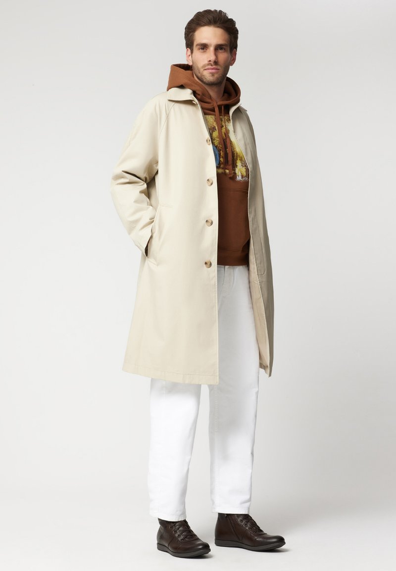 Beżowy trencz o prostym kroju, z dużymi guzami i bocznymi kieszeniami. Noszony na brązowej bluzie z kapturem, zestawiony z białymi spodniami i ciemnobrązowymi butami.