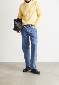 Homme portant un sweat à capuche jaune et un jean bleu, tenant un sac noir plié d'une main et l'autre main dans la poche, se tenant sur un sol gris.