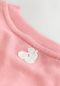 Roza bombažni pulover z rebranim izrezom in belim vezenim cvetličnim vzorcem na levi strani ovratnika.