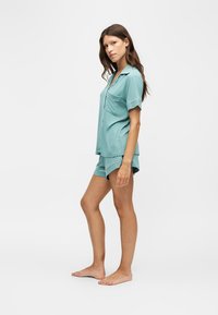 Femme debout pieds nus portant un haut boutonné bleu clair à manches courtes et un short de pyjama assorti sur un fond uni.