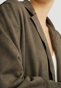 Chaqueta de ante marrón con cuello con muesca y textura suave, que presenta detalles de costura visibles y un ajuste holgado.