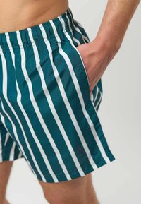 Homme portant un short rayé verticalement vert et blanc avec une taille élastique, main placée dans la poche droite, fond neutre.