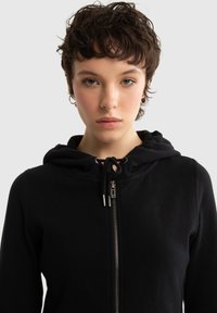 Młoda osoba z krótkimi kręconymi brązowymi włosami, ubrana w czarną bluzę z kapturem na zamek z troczkami, stojąca przodem na tle jednolitego jasnego tła.