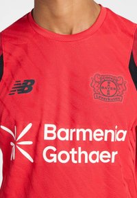 Camiseta deportiva roja con un patrón texturizado, detalles en negro, logo del club bordado y texto del patrocinador en blanco. Fabricada con tela transpirable.