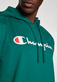 Zelená bezrukávová mikina s kapucí a šňůrkou, která má výrazné bílé logo "Champion" a malý červený akcent v písmenu "C".