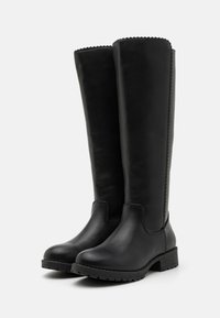 Bottes en cuir noir arrivant au genou, avec une texture lisse, un bout rond, un talon carré et un bord scallopé sur le haut. Semelle en caoutchouc pour la traction.