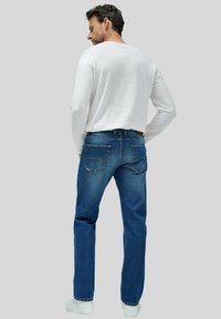 Vaqueros de mezclilla azul con corte recto, que cuentan con dos bolsillos traseros y un lavado medio. Combinados con una camisa blanca de manga larga y zapatillas deportivas.