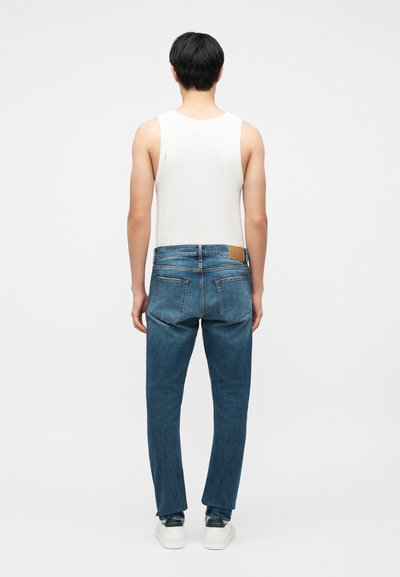 Άνδρας που φορά λευκό αμάνικο τοπ, μπλε τζιν και λευκά αθλητικά παπούτσια, στέκεται με την πλάτη προς το μέρος μας μπροστά σε απλό λευκό φόντο.